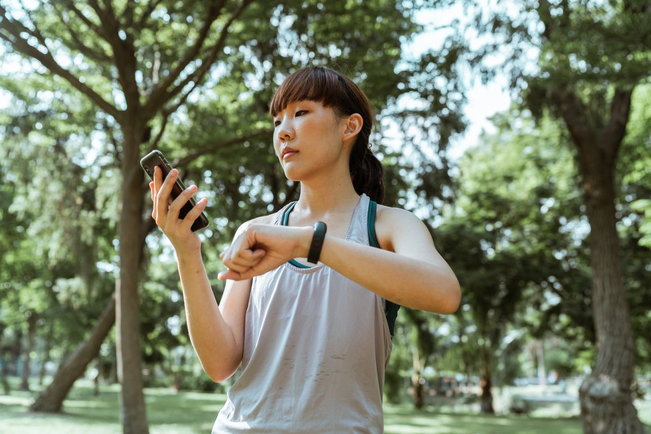 Mastering the First Impression: Your intriguing post title goes here Positive concentrated young fit Asian sportswoman in activewear browsing smartphone and checking fitness bracelet while training in green sunny park